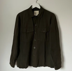Mörkgrön overshirt från A Day's March - Snygg mörkgrön overshirt från A Day's March i 100% bomull. Klassisk rak passform med två bröstfickor med knappstängning och knappar hela vägen framtill. Perfekt att slänga på över en t-shirt för en chill look. 5H9