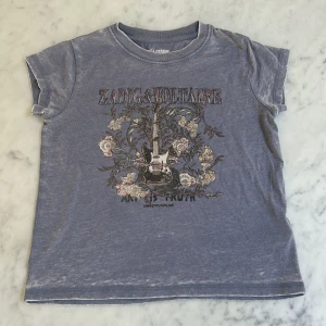 Blå t-shirt Zadig & Voltaire XS - Blå t-shirt från Zadig & Voltaire i storlek XS med coolt tryck av elgitarr och blommor på bröstet. T-shirten har korta ärmar, rund halsringning och är gjord i mjuk bomull. Perfekt för dig som gillar rockig stil med en twist.