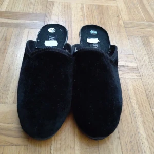 Svarta mockatofflor i storlek 37 - Svarta bekväma tofflor i mjuk mocka med platt sula och öppen häl. Perfekta att glida i hemma för en chill vibe. Snygg minimalistisk look och bekväm passform. Sula i syntetmaterial för extra hållbarhet. Har flera storlekar.