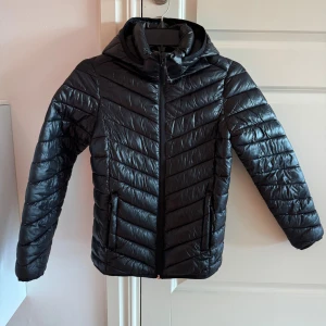 Svart dunjacka från McKinley - Svart dunjacka från McKinley med huva och dragkedja framtill. Passar höst eller lite mildare vinterdagar. Jackan har ett stilrent, quiltat mönster och två fickor med dragkedja. 