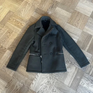 Armani Jacka - Stilren mörkgrön skepparkavaj från Armani Jeans i storlek 44 eller Small med klassisk dubbelknäppning, bred krage och två dragkedjeförsedda fickor. Jackan är fodrad med mjuk fleece och har både knappar och dragkedja framtill för extra edge. En knapp sitter ganska löst, men det går att använda dragkedjan bara, den går nog lätt att fixa själv eller så kan jag fixa den fär 100 extra.