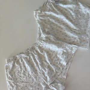 Blommigt pyjamaset från Kappahl - Supersöt linnetopp och shorts från Kappahl i ljusbeige med små blommor i rosa och blått. Tunt och mjukt bomullsmaterial med smala axelband och justerbar passform. Resor i midjan. Perfekt för varma dagar eller som mysig pyjamas.