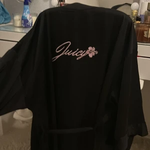 Svart kimono från Juicy Couture - Snygg svart kimono från Juicy Couture med broderad rosa logga och blomma på ryggen. Modellen har vida ärmar och knytskärp i midjan för en chill och lyxig vibe. Perfekt att slänga på sig hemma eller när du vill känna dig extra glam. Passar även dig som har storlek S/M