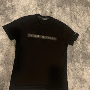 Svart Emporio Armani t-shirt M - Svart t-shirt från Emporio Armani med klassisk rund hals och korta ärmar. Framsidan har ett snyggt Emporio Armani-logotypband i svart och silver. Materialet är mjuk bomull som känns skön mot huden. Perfekt för dig som gillar stilrena designer och exklusiva märken.