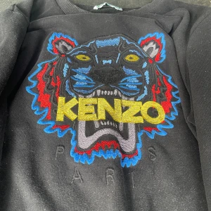 Svart sweatshirt från Kenzo med tiger - Cool svart sweatshirt från Kenzo med ikoniskt broderat tigermotiv i blått, rött, gult och grått på bröstet. Stort KENZO-tryck i gult och ribbade muddar vid hals, ärmslut och nederkant. Perfekt statement piece för din garderob. Pris kan diskuteras!