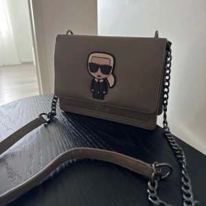 Beige axelväska Karl Lagerfeld - Snygg beige axelväska från Karl Lagerfeld med coolt motiv på framsidan. Väskan har en stilren rektangulär form, kedjerem i metall och är gjord i slätt skinn. Perfekt storlek för det viktigaste och har en modern vibe med signaturdetalj framtill.