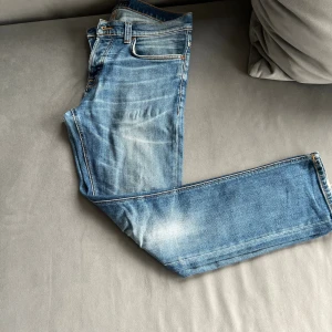 Blå jeans från Nudie med broderad ficka - Snygga blå jeans från Nudie med klassisk femficksdesign och coolt broderi på bakfickan. Jeansen har en rak passform och är tillverkade i mjukt denimtyg med slitningar och ljusa tvättade partier för en avslappnad look.