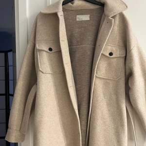 Beige overshirt från NLY Trend - Stilren beige overshirt från NLY Trend med klassisk krage, stora bröstfickor med knappar och rak passform. Jackan har långa ärmar och är tillverkad i ett mjukt material som ger en clean och modern look.