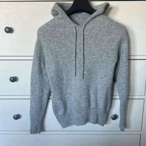 Grå stickad hoodie i kashmir - Mysig grå hoodie i kashmir med huva och snörning. Tröjan är stickad och har långa ärmar samt ribbade muddar vid ärmslut och nederkant. Perfekt för kyliga dagar och enkel att matcha med jeans eller mjukisbyxor.