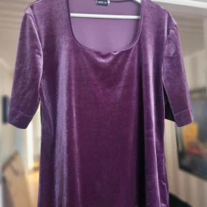 Lila sammetklänning från Add It - Säljer en lila topp i mjuk sammet från Add It. Klänningen har en snygg fyrkantig urringning och korta ärmar. Perfekt för dig som gillar en stilren look med lite lyxig känsla. Passar dig som vill sticka ut med färg och material.