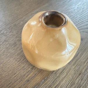 Ljusstake brun/beige - Bilderna visar en beige, glaserad ljusstake med oregelbunden form. Ytan är blank och ljusstaken ser ut att vara i gott skick utan synliga sprickor eller större skador. Perfekt som inredningsdetalj för hemmet.