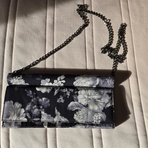Blommig kuvertväska från Whyred - Snygg kuvertväska från Whyred med blommigt mönster i djupblått och grått. Väskan har en silverfärgad kedja som axelrem och ett praktiskt fack med dragkedja inuti. Perfekt för att lyfta din outfit med lite extra stil.