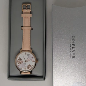 Blommig klocka från Oriflame - Snygg klocka från Oriflame med ljusbeige skinnarmband och rund urtavla i guldton. Urtavlan är vit med blommigt mönster i rosa och guld, samt stilrena romerska siffror. Perfekt accessoar för dig som gillar detaljer och stilren design.