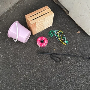 Trälåda,plasthink,hönät,spö,rosettband - Bilderna visar en rosa plasthink, en trälåda, ett band på rulle, ett färgglatt nät och en svart pinne. Inga elektronikprodukter syns på bilderna, endast hushålls- och hobbyartiklar. 