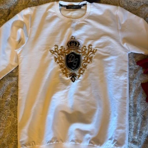 Vit sweatshirt från Dolce & Gabbana - Säljer en vit sweatshirt från Dolce & Gabbana med ett stort broderat emblem i svart och guld på bröstet. Tröjan har rund halsringning och långa ärmar. Perfekt för dig som vill sticka ut med en lyxig och unik look.