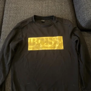 Hugo boss tröja  - Svart långärmad tröja från Hugo Boss med stort, glansigt guldtryck på bröstet. Köpt från Zalando.  Klassisk rund halsringning och stilren design som sticker ut med den feta loggan framtill. Perfekt för dig som gillar streetstyle och vill ha något som syns. 