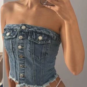 Blå denim tubtopp med knappar - Säljer en snygg tubtopp i blå denim med fransig nederkant och knappar framtill. Toppen har två bröstfickor med lock och elastisk smock baktill för skön passform. Perfekt för dig som gillar en edgy och trendig stil.
