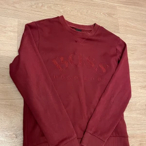 Vinröd sweatshirt från Hugo Boss - Säljer en vinröd sweatshirt från Hugo Boss med broderad logga framtill. Tröjan har rund hals, långa ärmar och ribbade muddar vid ärmslut och nederkant. Perfekt för dig som gillar stilren och klassisk streetwear.