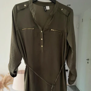 Olivgrön kortklänning från H&M - Olivgrön kortklänning från Divided med långa ärmar som kan rullas upp och fästas med knapp. Klänningen har v-ringad hals, guldfärgade knappar och dragkedjor vid bröstet samt ett knytband i midjan för snygg passform.