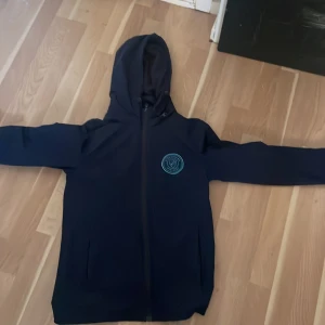 Svart hoodie med Djurgården märket på bröstet  - Det är en Svart hoodie som är stor i storlek den sitter som en XS DEN har hel dragkedja och huva. På bröstet finns ett blått, runt tryck med märke av Djurgården och emblem. Tröjan har långa ärmar och två fickor framtill. Perfekt för chill dagar eller när du vill ha en enkel streetstyle-look.