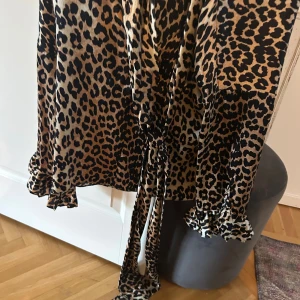 Leopardmönstrad omlottklänning Ganni - Snygg leopardmönstrad omlottklänning från Ganni i lyxig silkesblandning. Klänningen har långa ärmar med volangdetaljer vid ärmslut och knytband i midjan. Färgerna går i beige och svart, perfekt för dig som vill sticka ut med en trendig look.