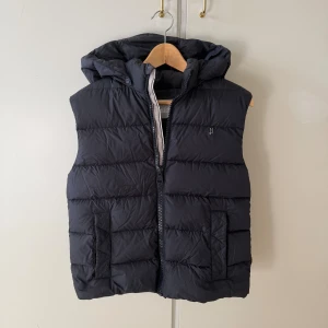 Svart dunväst från Herno med huva - Mörk blå quiltad dunväst från Herno med avtagbar huva och dragkedja framtill. Västen har två stora fickor och diskret H-logo på bröstet. Perfekt för lager-på-lager och snygg till både hoodie och t-shirt. Vill bara bli av med den därför så bra pris 