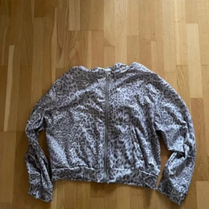 Grå leopardmönstrad hoodie - Säljer en grå hoodiejacka med leopardmönster och dragkedja framtill. Jackan har huva, långa ärmar och två fickor. Perfekt för dig som gillar streetstyle och vill sticka ut med ett djurmönster.