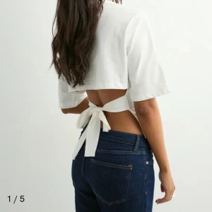Nelly tie tee - Säljer en vit croppad topp med korta ärmar och öppen rygg som knyts med en bred rosett baktill. Toppen har en stilren och modern look, perfekt för dig som gillar trendiga detaljer och vill visa lite hud. Materialet känns lätt och passar bra till jeans.
