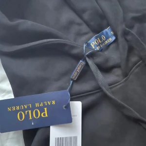 Svart hoodie från Polo Ralph Lauren - Svart hoodie från Polo Ralph Lauren med klassisk röd liga. Tröjan har huva med snörning och en stor magficka framtill. Tillverkad i mjuk bomull, perfekt för en chill och stilren look.