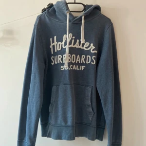 Mörkblå Hollister hoodie med tryck - Mörkblå hoodie från Hollister med vit text 'Hollister Surfboards SO. CALIF' på bröstet. Tröjan har huva med vit snörning och en klassisk känguruficka framtill. Perfekt för en avslappnad och cool stil. Storlek L men sitter som M