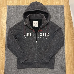 Fodrad Hollister Kofta - Mysig grå hoodie från Hollister med vit och röd logga på bröstet. Tröjan har dragkedja, huva och svart teddyfoder på insidan. Perfekt för kyliga dagar och har två fickor framtill. (liten slitning/hål på vänsta ärmen, men är verkligen inget man lägger märke till alls!) Hör av vid frågor, priset ej hugget i sten!!