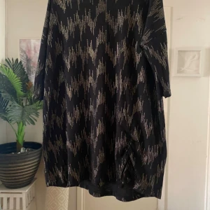 Svart glittrig kortklänning/tunika med guldmönster - Svart kortklänning med glittrigt guldmönster och snygg rynkdetalj på sidan. Klänningen har halvkort ärm och är tillverkad i ett stretchigt material som ger en bekväm passform. Perfekt för dig som vill sticka ut med en cool och trendig look.