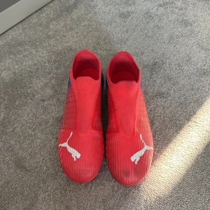 Röda Puma fotbollsskor strl 38,5 - Säljer ett par snygga röda Puma fotbollsskor med blå detaljer och vit logga. Skorna har en strömlinjeformad design utan snörning och är tillverkade i syntetmaterial med texten 'GripControl Speedcage' på sidan. Perfekta för dig som vill sticka ut på planen. Dem är använda under 3 månader. Köper dem för 1000 kr 