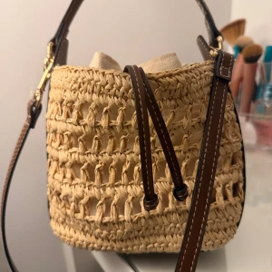Beige axelväska i flätad strå - Snygg axelväska i beige flätad strå med bruna detaljer och justerbar axelrem. Väskan har en bucket-form och stängs med snörning upptill. Perfekt för en chill sommarlook och rymmer det viktigaste du behöver ha med dig.