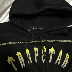 Trapstar svart hoodie med tryck - Svart hoodie från Trapstar med huva och snörning. Framsidan har ett stort Trapstar-tryck i gul och grå gradient. Kontraststickningar längs ärmar och bröst ger en cool detalj. Perfekt för dig som gillar streetwear och vill sticka ut.