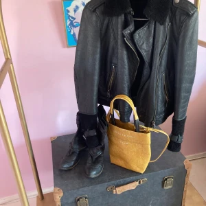 Svart Shearlingjack med fårskinn - Säljer en svart skinnjacka med fårskinn vid krage och ärmslut. Jackan har coola detaljer som dragkedjor framtill och på fickorna, samt spännen vid handlederna. Perfekt för dig som gillar en edgy stil och vill sticka ut.