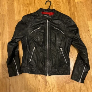 Svart skinnjacka Rockandblue 36 - Snygg svart skinnjacka från Rockandblue i storlek 36. Jackan har coola silverfärgade dragkedjor på fickor och ärmslut, quiltade detaljer på axlarna och röd insida. Figursydd modell med ståkrage och knapp. Perfekt för dig som vill ha en edgy look.