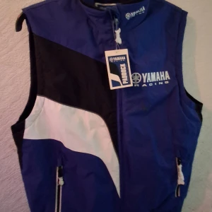 Yamaha Racing blå dunväst - Sportig blå dunväst från Yamaha Racing med vita och svarta detaljer, dragkedja framtill och två fickor med dragkedja. Insidan är quiltad och fodrad med blå fleece för extra komfort. Perfekt för dig som gillar racing och vill sticka ut.