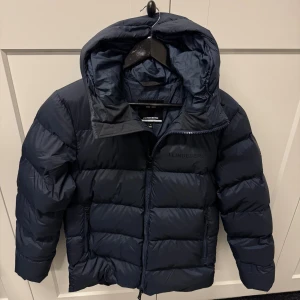 Mörkblå dunjacka från J.Lindeberg - Snygg mörkblå dunjacka från J.Lindeberg med huva och dragkedja framtill. Jackan har en puffig design, två sidofickor med dragkedja och diskret logga på bröstet. Perfekt för kalla dagar när du vill hålla stilen och värmen.