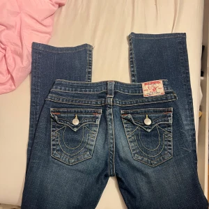 True Religion blå jeans - Säljer ett par klassiska blå jeans från True Religion med ikoniska kontrastsömmar och snygga fickdetaljer med silverknappar. Modellen har låg midja och bootcut, samt tydliga logopatch bak. Perfekt för dig som gillar street och premium denim.