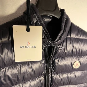 Mörkblå väst från Moncler - Snygg mörkblå glansig dunväst från Moncler med klassisk logga på bröstet. Västen har hög två fickor med dragkedja. Storlek är 3 vilket motsvarar S/M. Otroligt bra skick utan några skador, dragkedjan är dock utbytt professionellt för att dragkedjan på denna modell inte brukar vara så högkvalitativ. Säljer för att den har blivit för liten. västen är helt äkta och kostar upp mot 9000kr ny!