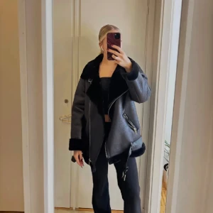 Svart pälsjacka med bälte och dragkedjor - Säljer en oversized svart pälsjacka med bred krage, bälte i midjan och detaljerade dragkedjor. Jackan har långärmade ärmar och mjukt foder, perfekt för kalla dagar. Snygg och trendig modell som ger en cool vibe.