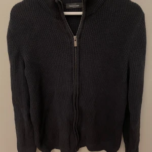 Svart stickad kofta från Dressmann - Säljer en svart stickad kofta från Dressmann med hel dragkedja framtill och ribbad krage. Perfekt för lager-på-lager och passar till många olika stilar. Klassisk och enkel design som funkar året runt.