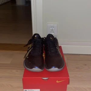 Nike Kobe 5 Protro svart/guld sneakers - Säljer ett par Nike Kobe 5 Protro sneakers i svart med metallic guld-detaljer och svart snörning. Skorna har en låg silhuett, perforerad tå och en cool guldmönstrad swoosh på sidan. Kommer med originalröd box och exklusiv röd dustbag med Kobe-logga.