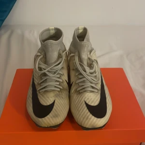 Nike Mercurial beige fotbollsskor - Nike Mercurial fotbollsskor i ljusbeige och med svarta detaljer och stor Swoosh-logga på sidan. Skorna har snörning, platt sula med dobbar och en strumpliknande hög krage för extra stöd. Lite smutsiga men funkar väldigt bra ändå.