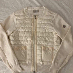 Vit quiltad cardigan jacka från Moncler - Snygg vit cardigan jacka från Moncler med quiltad framsida och två fickor med tryckknappar. Jackan har ribbade muddar, dragkedja framtill och Moncler-logga på ärmen. Materialmix med dunfylld front och stickade ärmar ger en exklusiv och sportig look. Har prislappen kvar