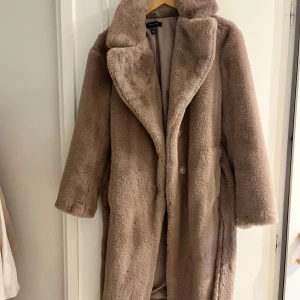 Lila pälsjacka med bälte från New Look - Supermjuk och fluffig ljuslila pälsjacka från New Look. Jackan har bred krage, knytbälte i midjan och långa ärmar. Perfekt för att hålla sig varm och snygg under kyliga dagar. Fodrad insida för extra komfort.