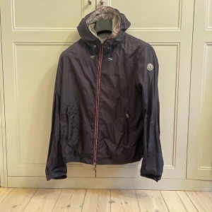 Moncler jacka - Snygg mörkblå vindjacka från Moncler med huva och klassisk logga på ärmen. Jackan har dragkedja med röd, vit och blå kant, två fickor med dragkedja och justerbar nederkant. Perfekt för dig som gillar stilren design och premiumkänsla. Såklart äkta!storlek 3 motsvarar medium 