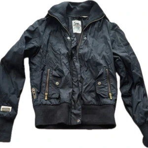 Svart bomberjacka från Soulstar - Svart bomberjacka från Soulstar med ribbade muddar och krage, dragkedja framtill och två fickor med dragkedja och tryckknapp. Jackan passar storlek S. Materialet är polyester med detaljer i tyg och metall. Endast använd 1 gång. 