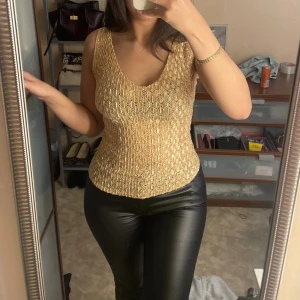Glittrig guldig ärmlös topp - Supersnygg ärmlös topp i glittrigt guldigt tyg med strukturerad yta. Toppen har v-ringning och croppad passform, perfekt för att sticka ut. Materialet är glansigt och känns lyxigt, med en cool textur som fångar ljuset.